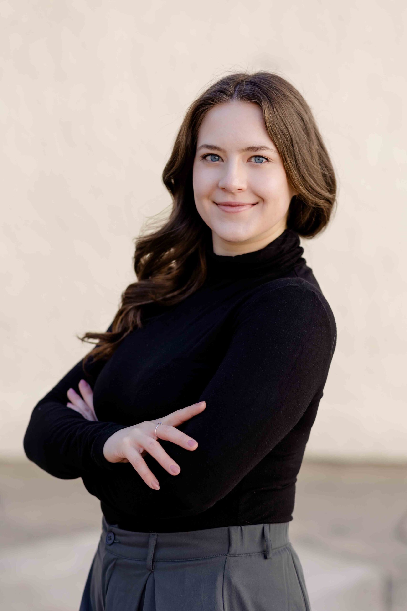 Authentisches Bewerbungsfoto einer jungen Frau mit leichtem Lächeln, verschränkten Armen, schwarzem Oberteil und neutralem Hintergrund – professionell und natürlich aufgenommen - Bewerbungsfotos Freiberg