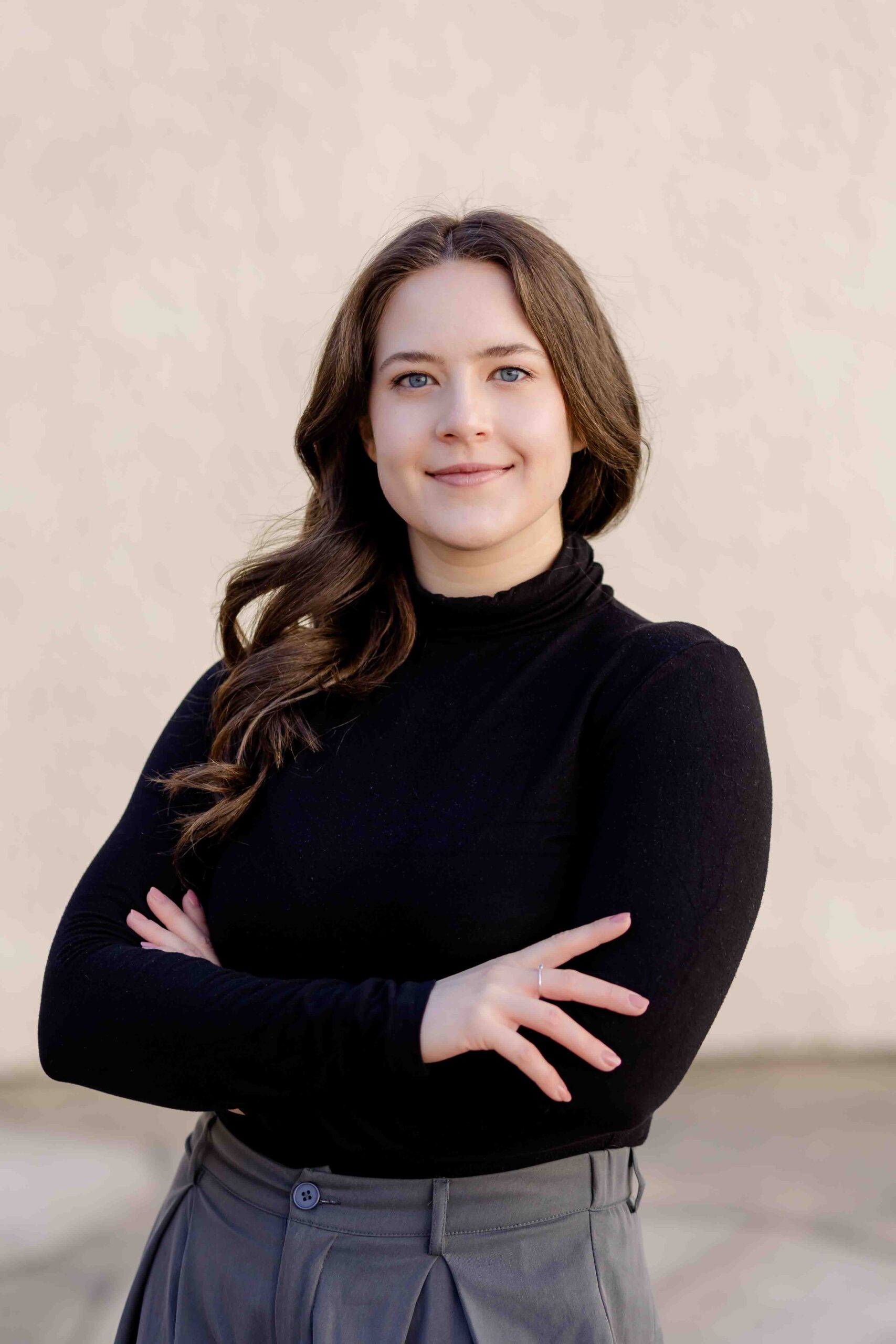 Authentisches Bewerbungsfoto einer jungen Frau mit leichtem Lächeln, verschränkten Armen, schwarzem Oberteil und neutralem Hintergrund – professionell und natürlich aufgenommen - Bewerbungsbilder Freiberg