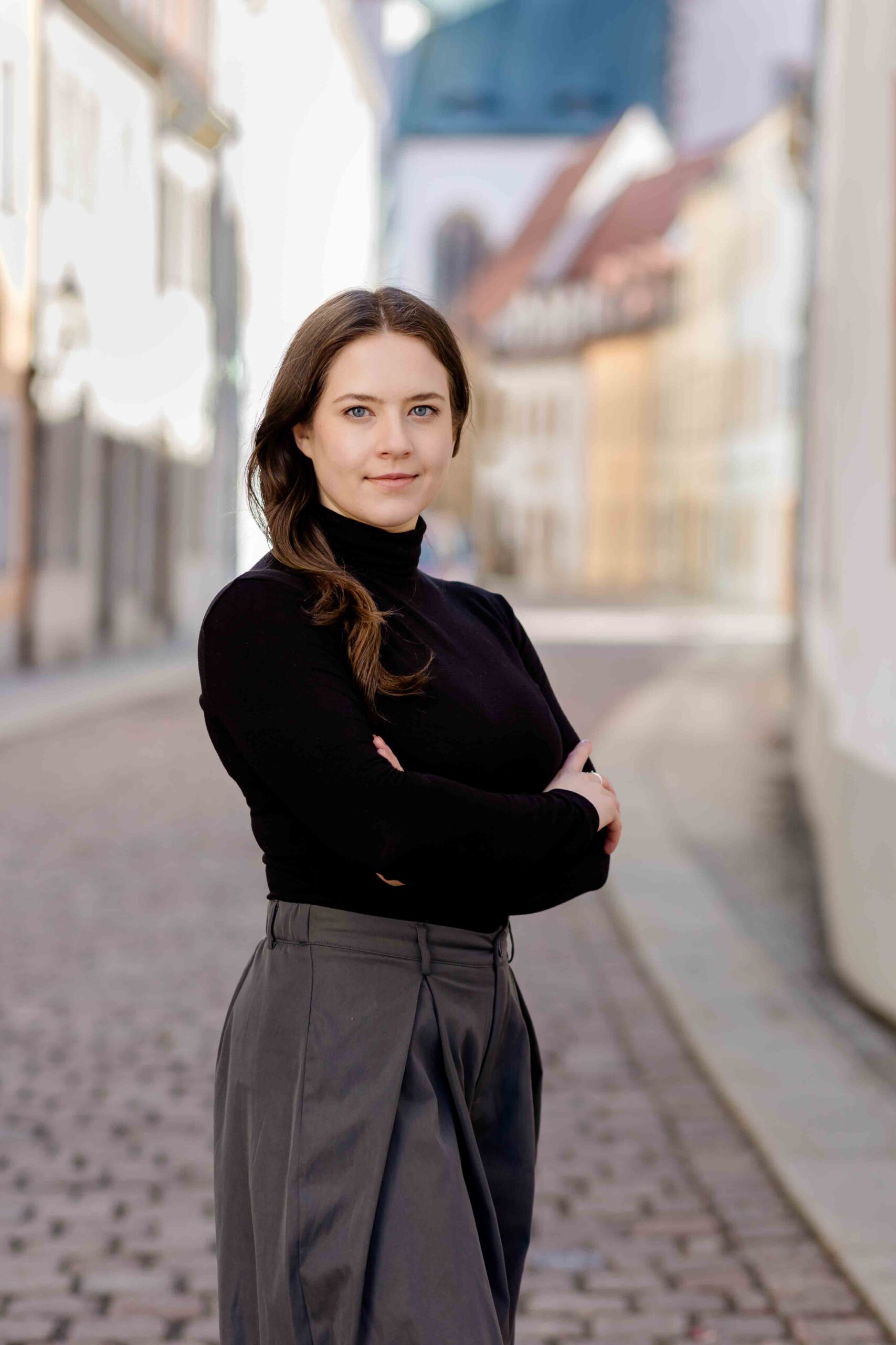 Authentisches Bewerbungsfoto einer jungen Frau mit leichtem Lächeln, verschränkten Armen, schwarzem Oberteil und neutralem Hintergrund – professionell und natürlich aufgenommen - Bewerbungsfotos Freiberg