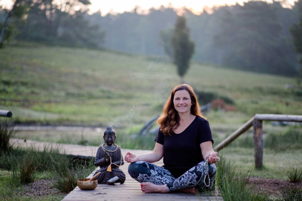 Businessfotos Yoga: Yogalehrerin sitzt meditierend im Schneidersitz auf einem Holzsteg in der Natur, ruhige Abendstimmung mit Buddhafigur im Vordergrund